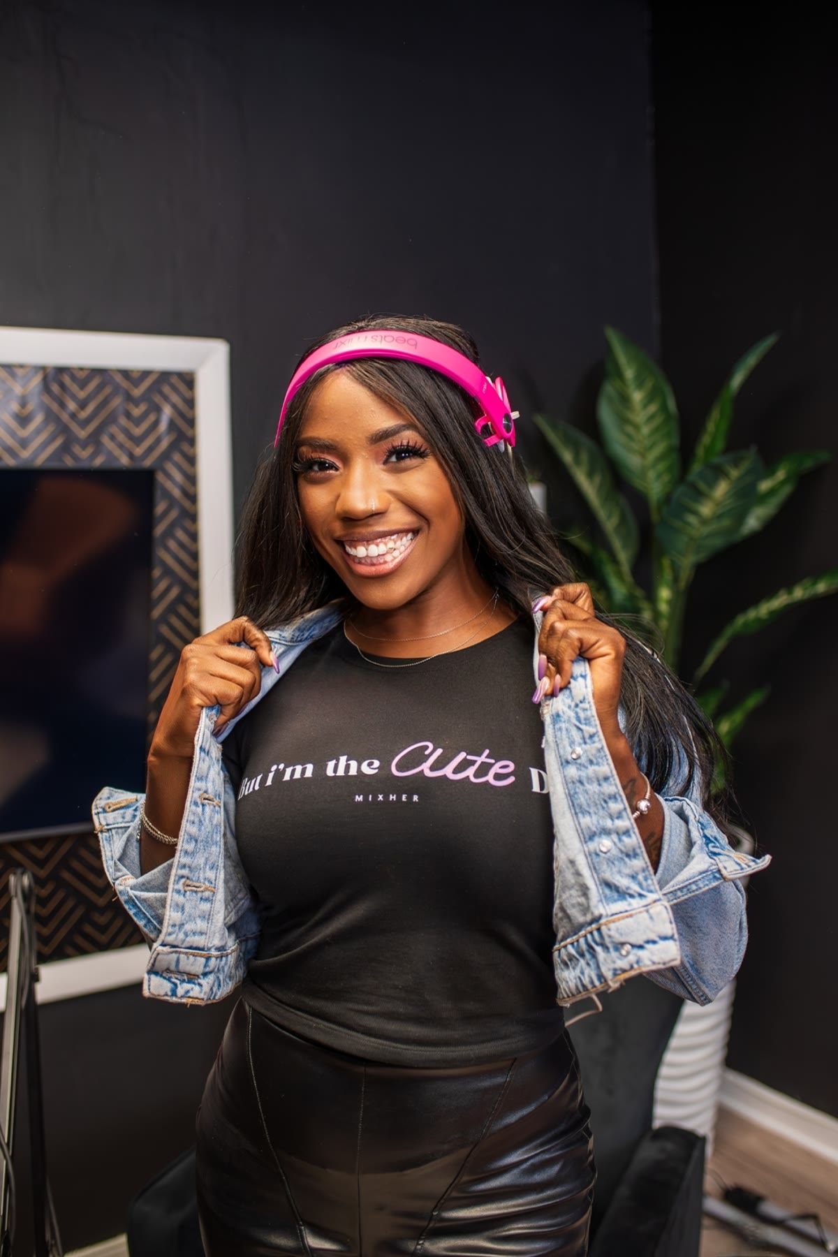 DJ ReRe, founder of First Spin, smiling in her studio with pink headphones and a denim jacket.